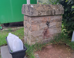 Small brick structure with a water tap attached. A small container is next tot he structure with old, empty milk bottles inside.